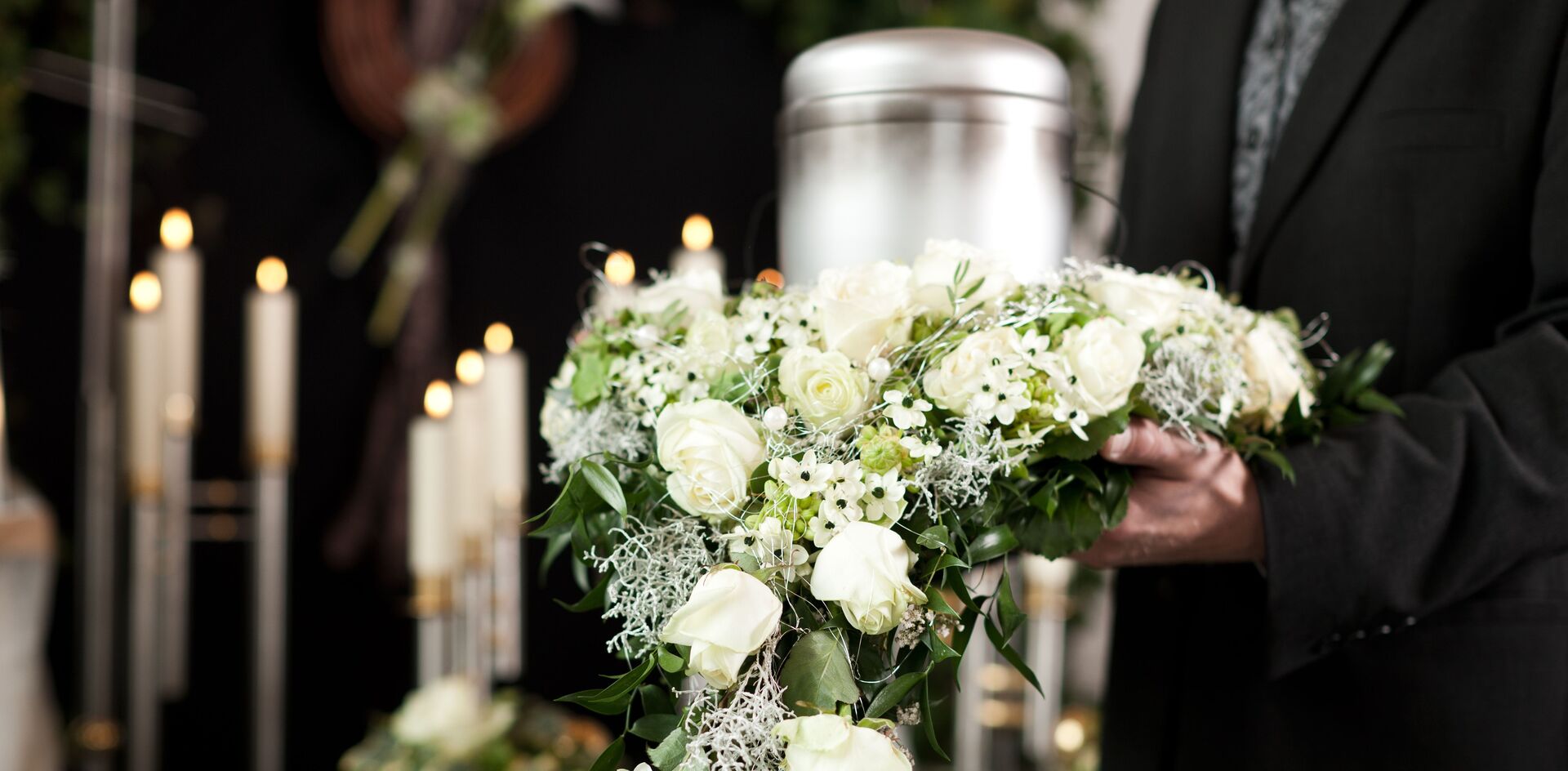 Eine Person in schwarzer Kleidung hält ein weißes Blumenarrangement bei einer Beerdigung mit einer Metallurne und Kerzen im Hintergrund.