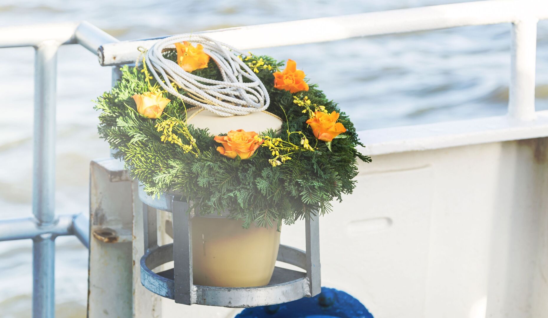 Blumenkranz mit orangefarbenen Blumen und Seil an einer Bootsreling, Wasser im Hintergrund.